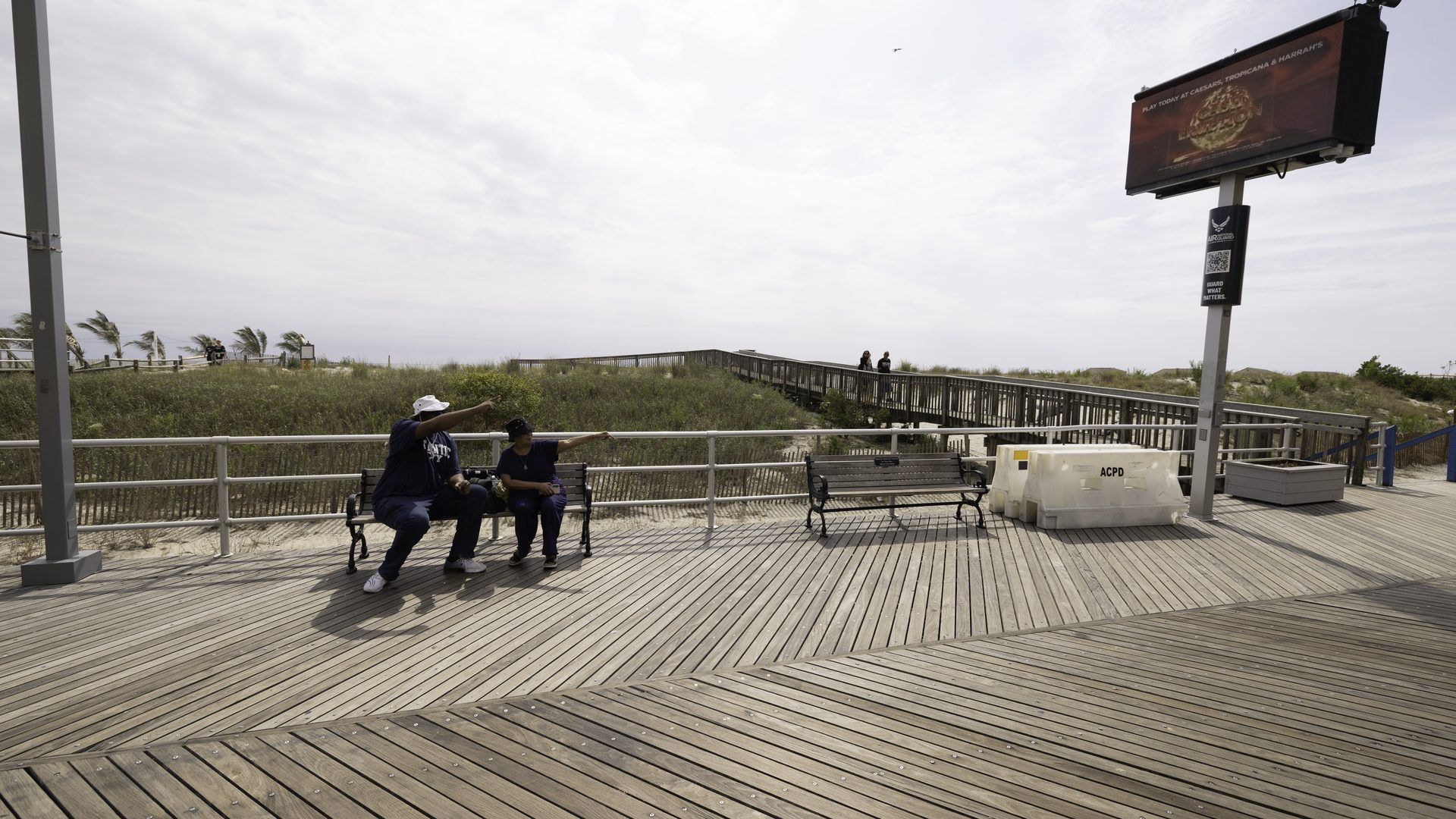 Two people on a Boardwalk bench, one pointing up at an APM digital screen