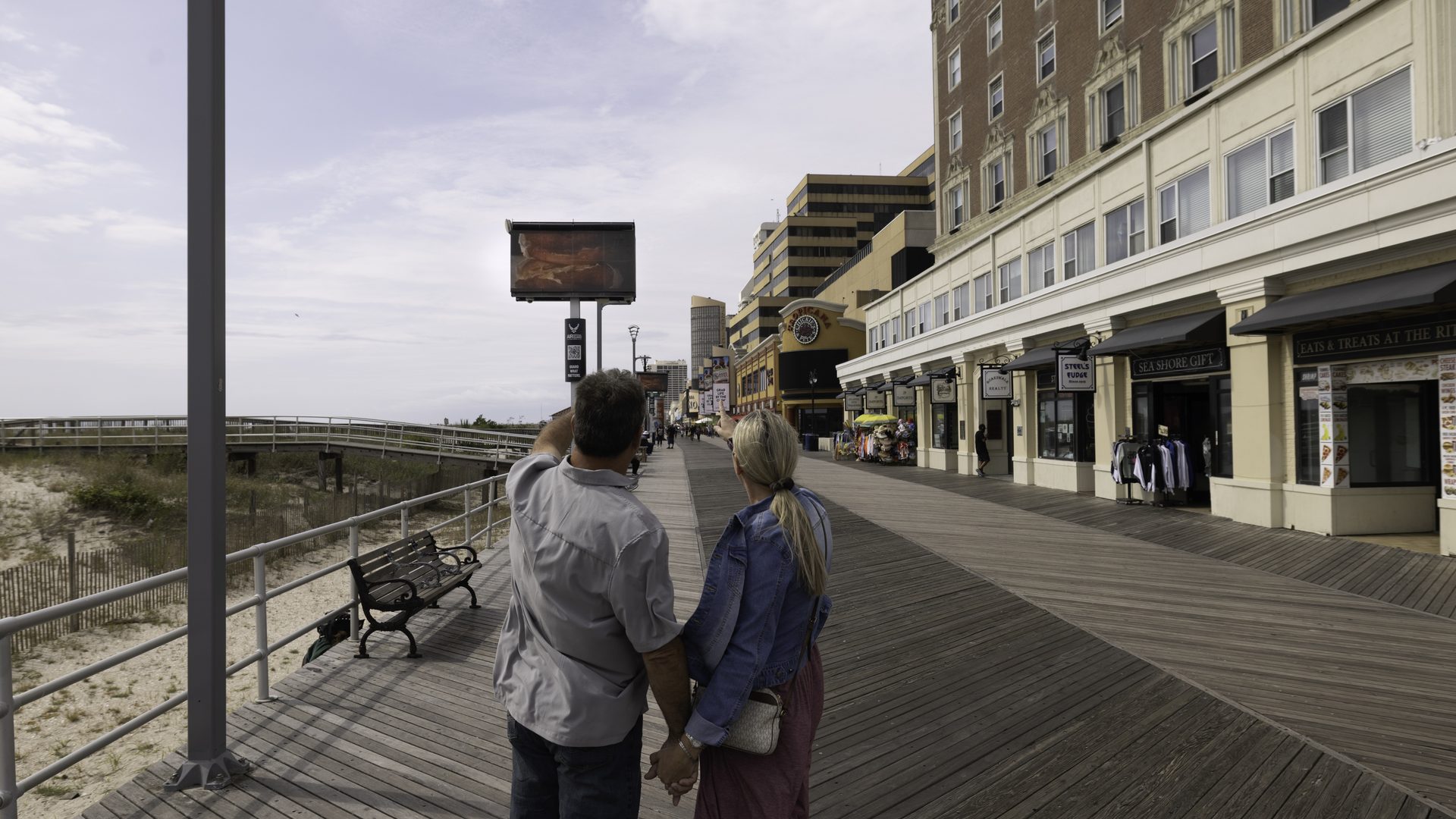 A couple walking hand in hand toward an APM digital screen on the Boardwalk, beach and dunes to the left