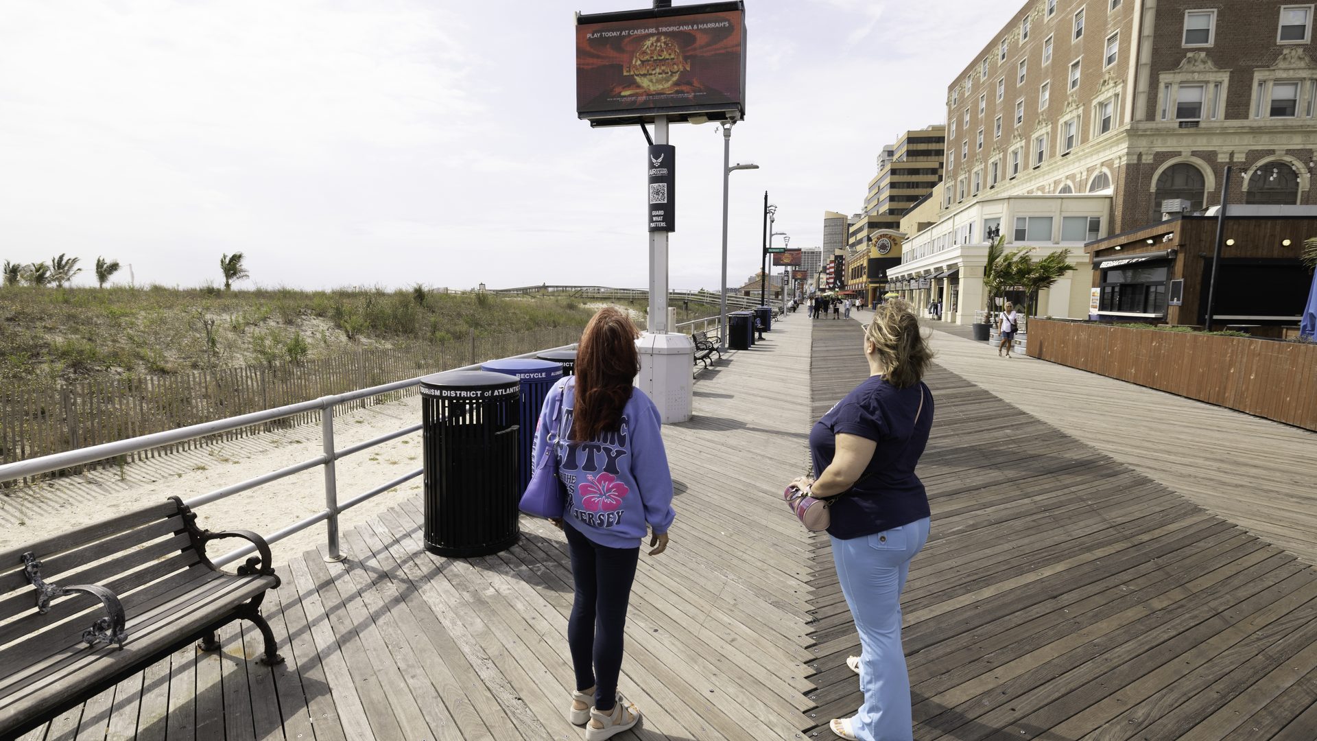 Two women walking the Atlantic City Boardwalk past an APM digital screen, hotels and restaurants along the right